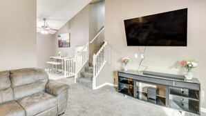 Sala de estar com sofá cinza, televisão de tela plana na parede, escada e uma pequena mesa com flores.