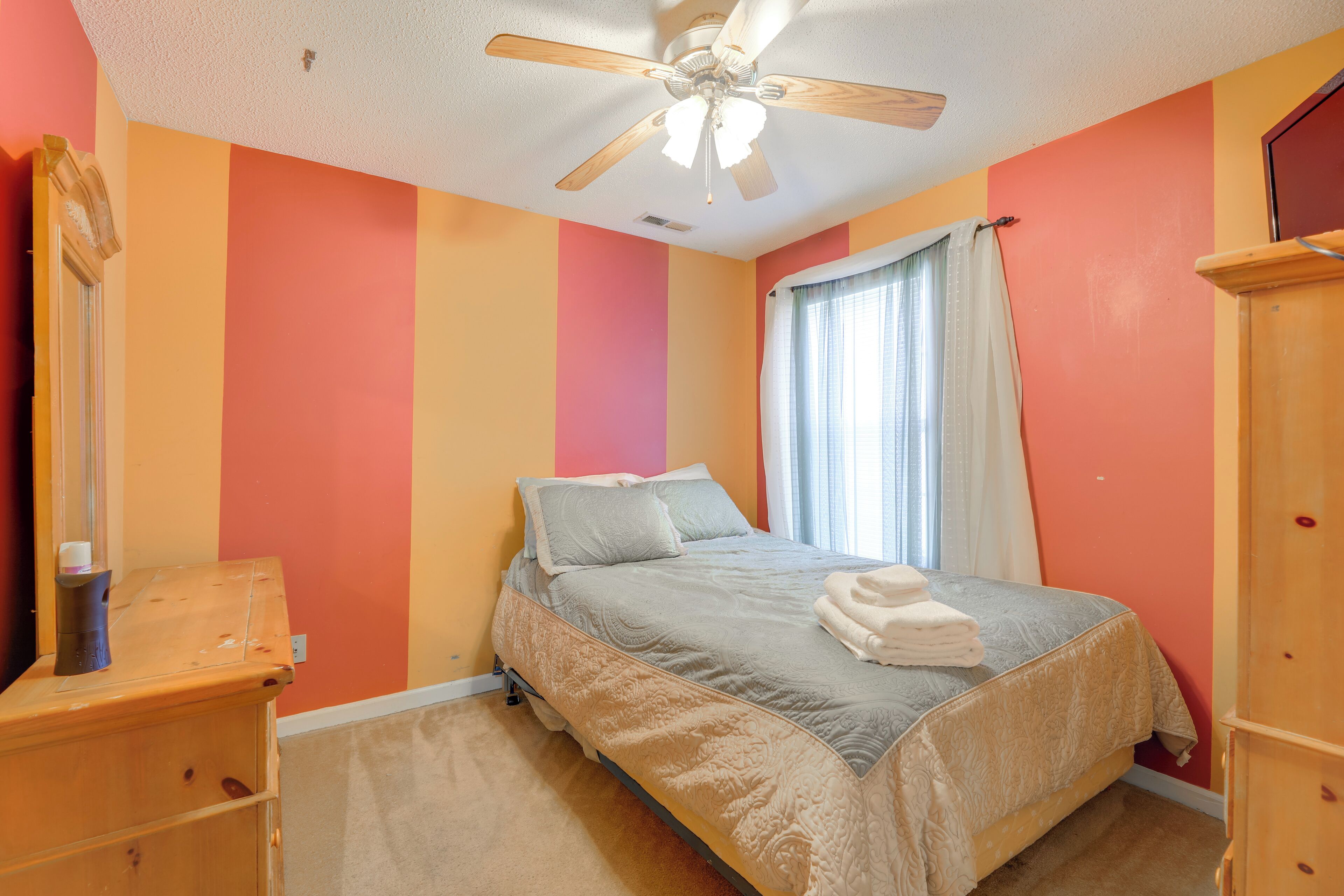A bedroom with a bed, a ceiling fan, a wooden dresser, and a window with curtains.