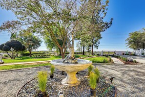 Un jardin avec une fontaine centrale, entouré de gravier, de plantes en pot et d’arbres.
