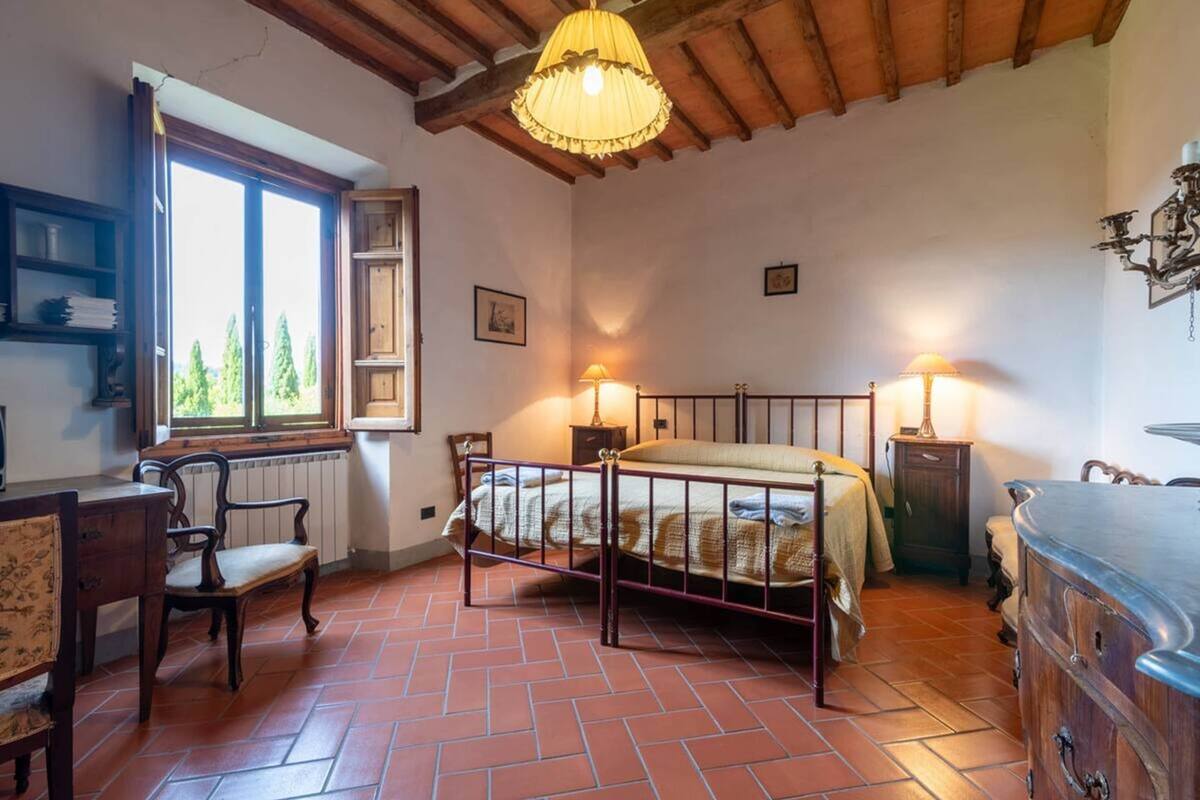 A bedroom with a bed, two bedside lamps, a wooden chair, a dresser, and a window with a view of trees.