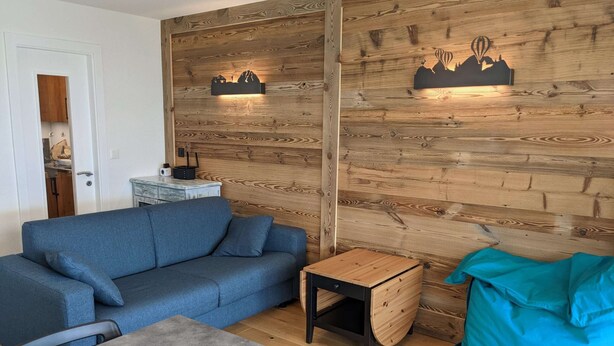 A room with a blue sofa, a small wooden table, a teal bean bag, and a wall with wooden paneling and two illuminated wall art pieces. - Cosy Apartment in a Central Location (Veysonnaz)