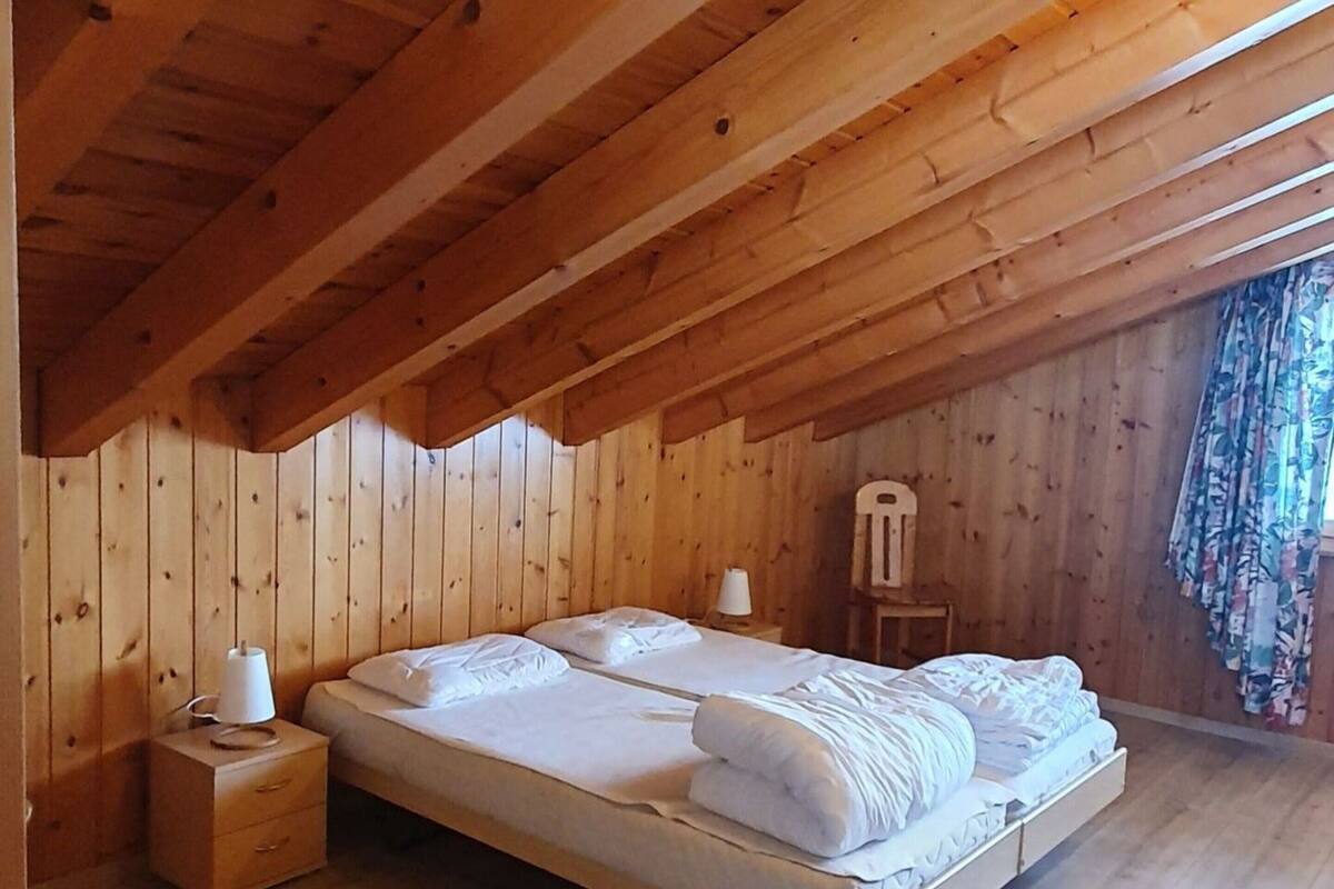 A single-bed room with wooden walls and ceiling, a nightstand with a lamp, and a chair.