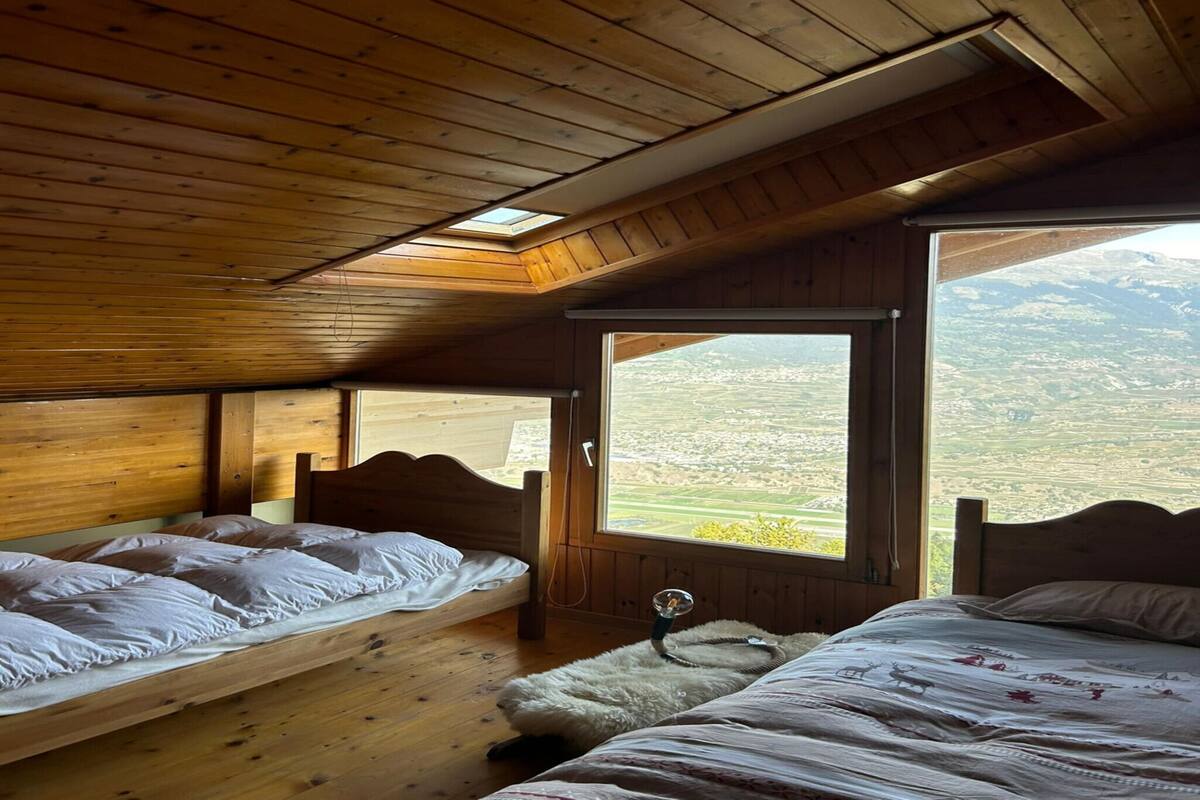 A wooden cabin with two beds, a skylight, and a large window offering a view of the countryside.