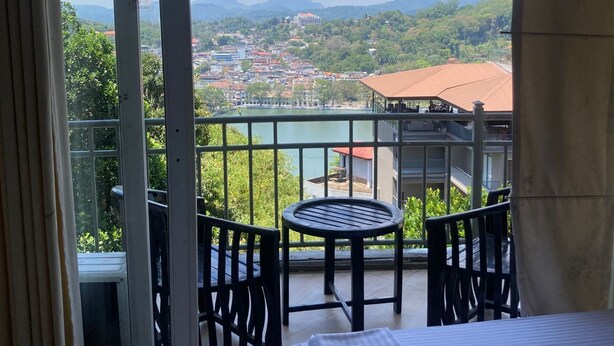 A balcony with a bed, a table, and chairs, overlooking a cityscape with mountains. - Hotel Sandriana Kandy (Kandy)
