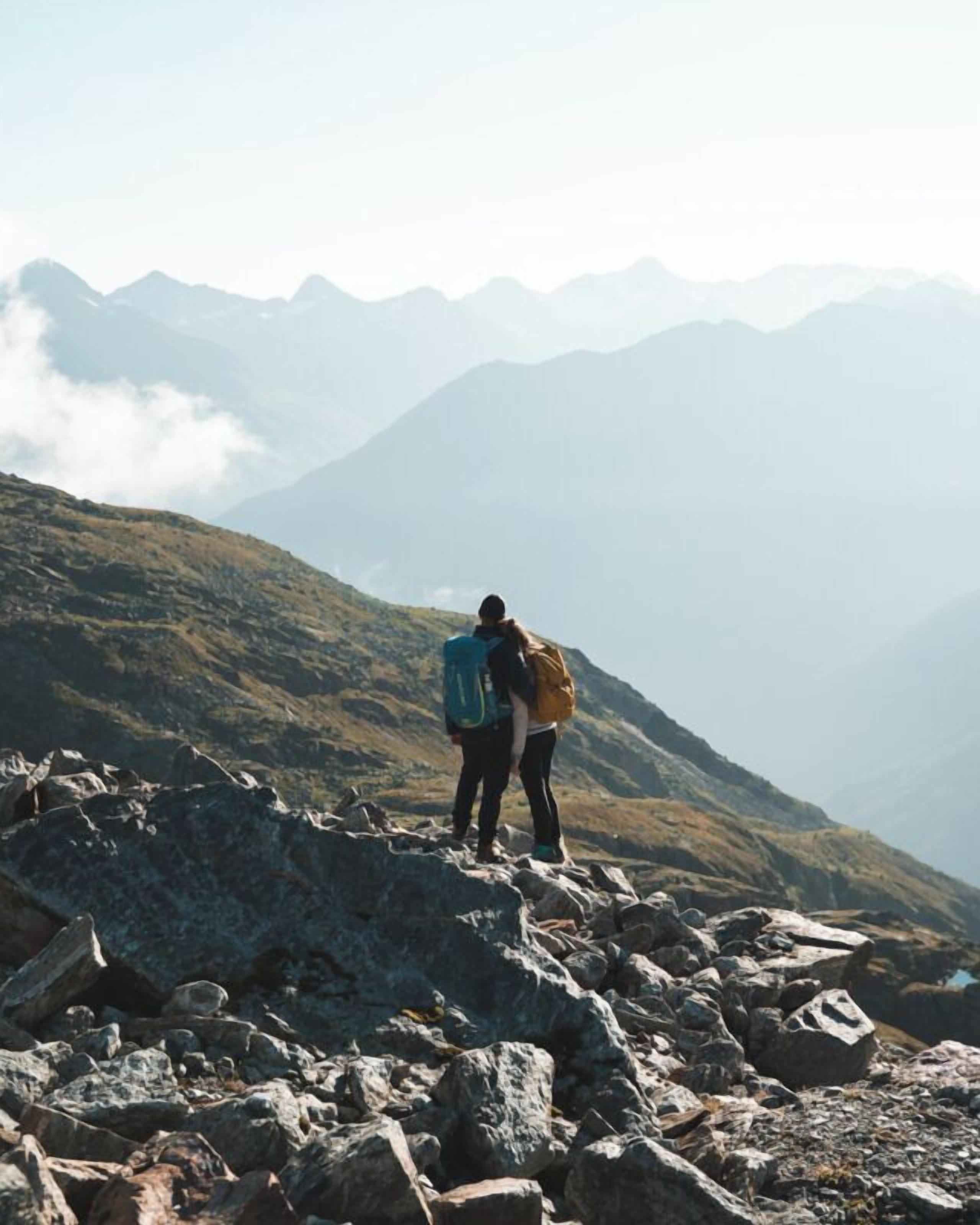 Dos personas caminan por un sendero pedregoso en la montaña, con vistas a montañas distantes.