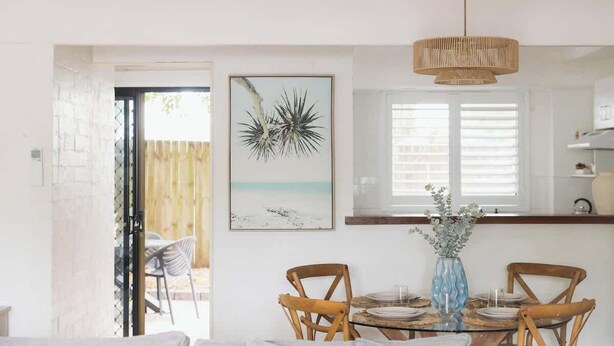 A dining area with a round table, chairs, a vase with flowers, and a beach-themed painting on the wall. - A Noosa Gem on Noosa Parade - close to everything (Noosa Heads)