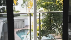 A view of a swimming pool with a wooden deck, surrounded by palm trees and a white building in the background. - A Noosa Gem on Noosa Parade - close to everything (Noosa Heads)