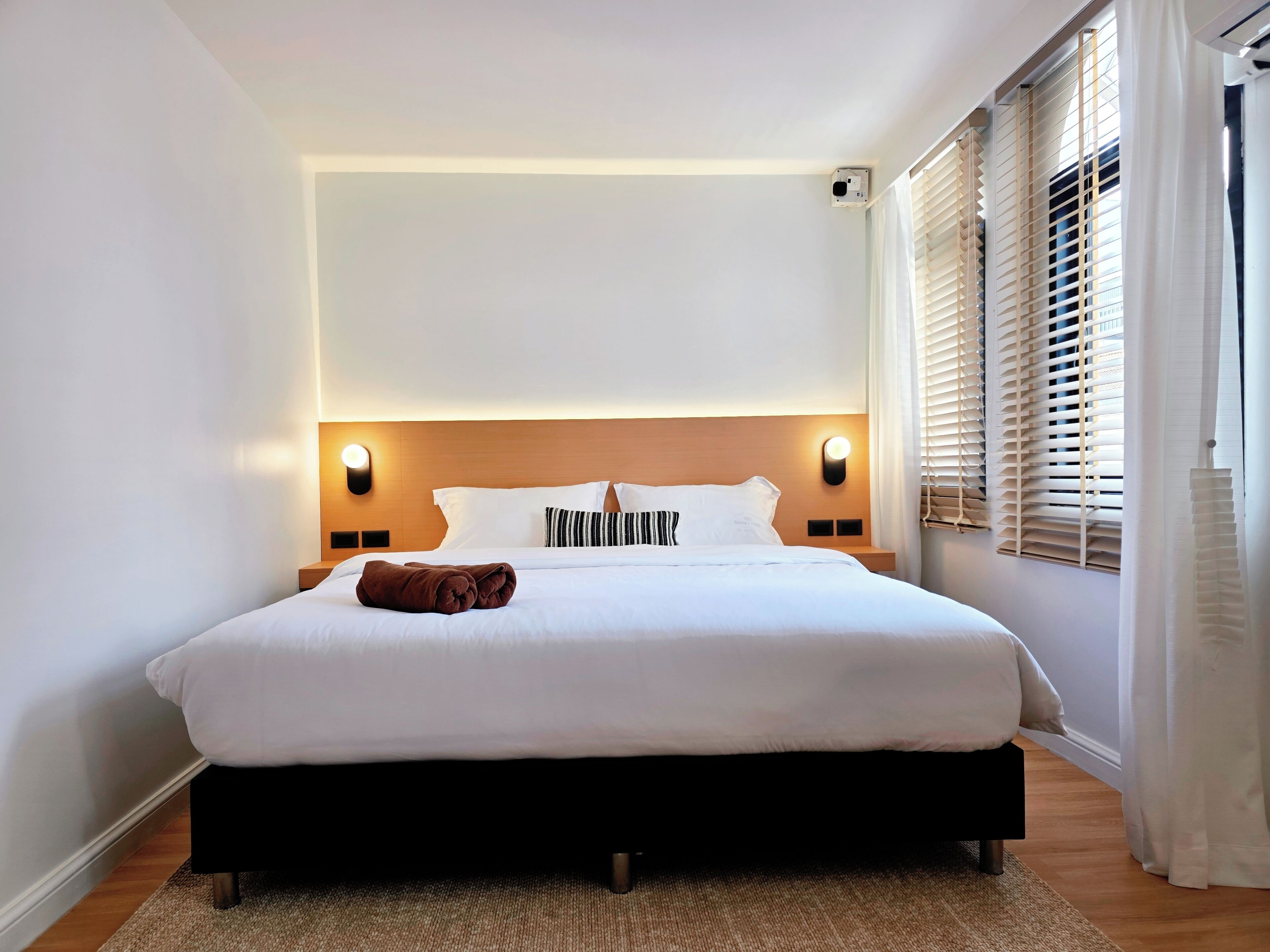 A hotel room with a large bed, two bedside tables, a headboard with integrated lighting, a window with blinds, and a wall-mounted air conditioning unit.