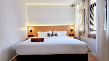 A hotel room with a large bed, two bedside tables, a headboard with integrated lighting, a window with blinds, and a wall-mounted air conditioning unit.