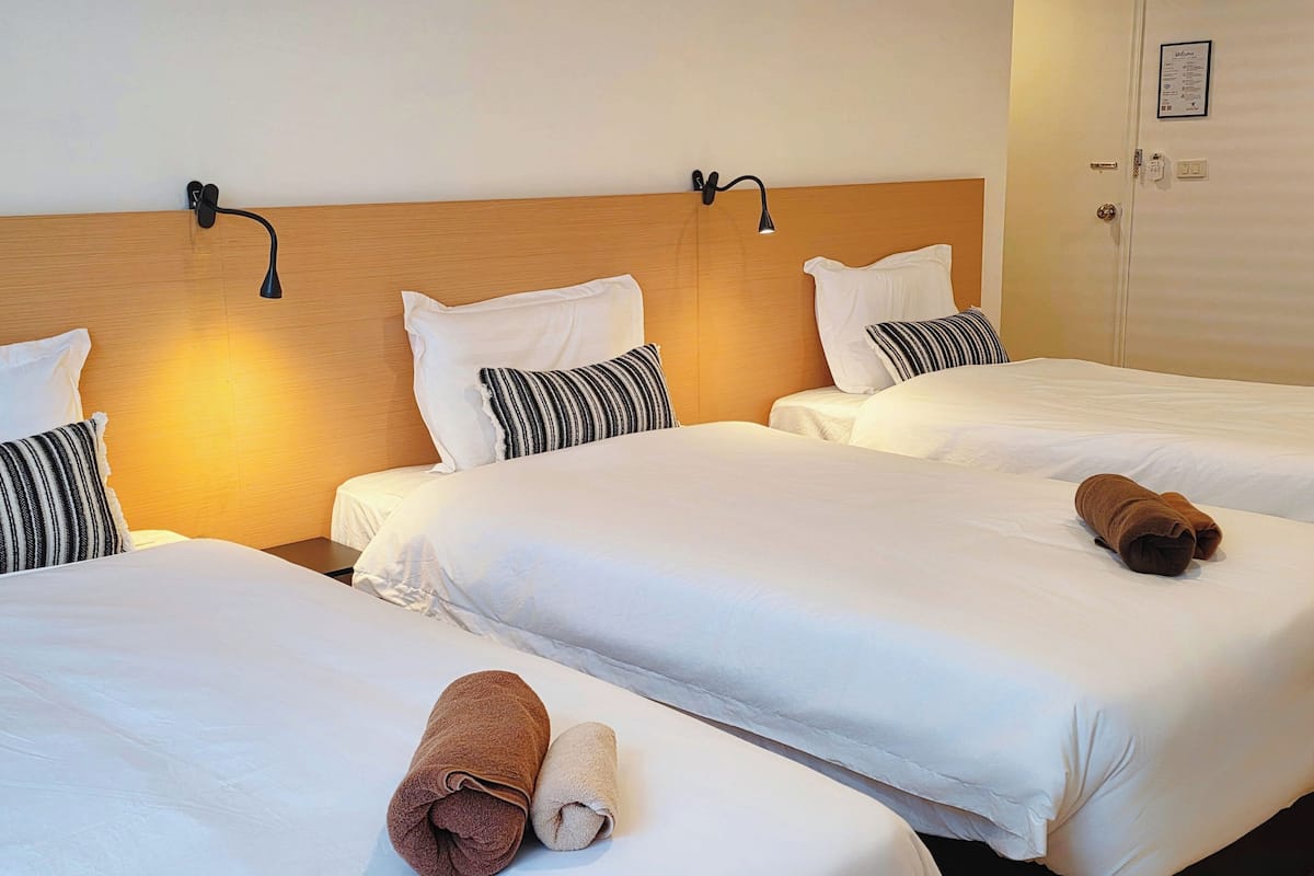 A hotel room with three beds, each with white linens and striped pillows. There are two wall-mounted lamps on the headboard.