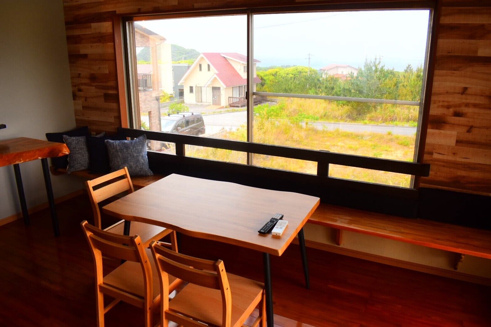 Um quarto com uma mesa e cadeiras de madeira, um banco e uma janela grande com vista para casas e vegetação.