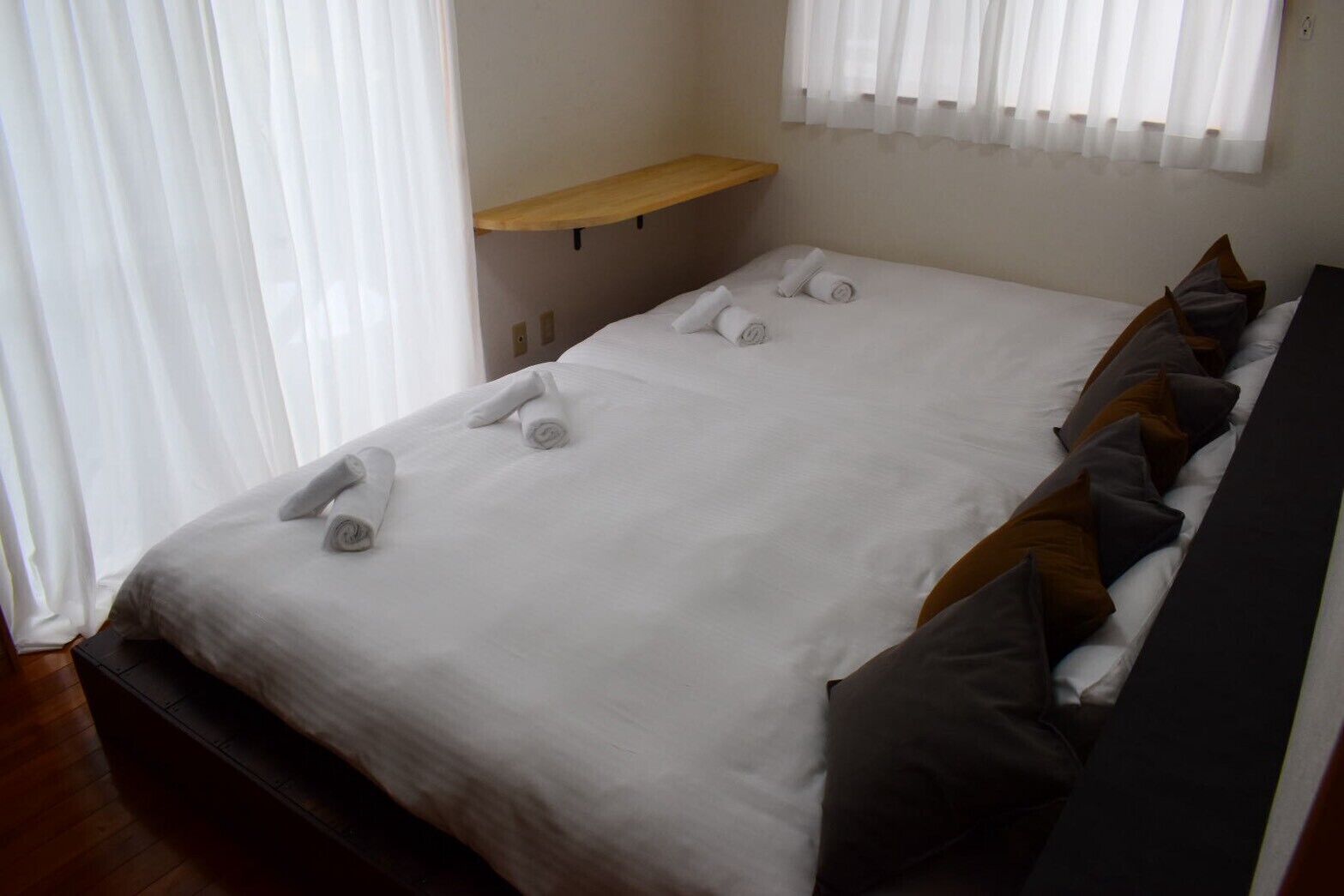 A double bed with white linens and rolled towels, a wooden shelf, and a window with white curtains.