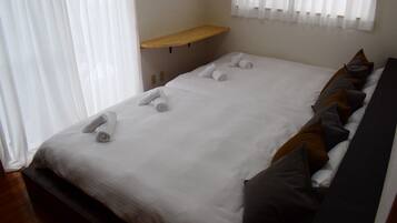 A double bed with white linens and rolled towels, a wooden shelf, and a window with white curtains.