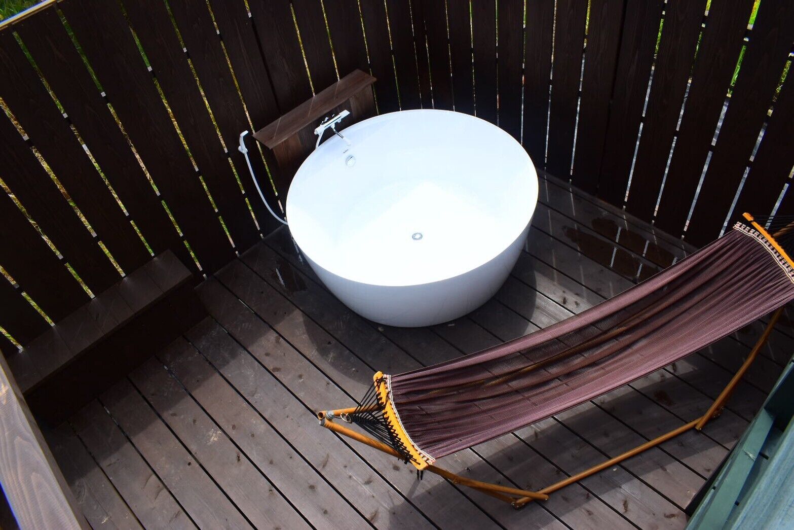 A wooden deck with a round bathtub, a hammock, and a small wooden stool.