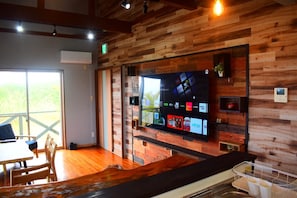 Ein modernes Interieur mit einer Holzverkleidung, einem an der Wand montierten Fernseher, der verschiedene Sender zeigt, und einer Essecke mit Tisch und Stühlen.