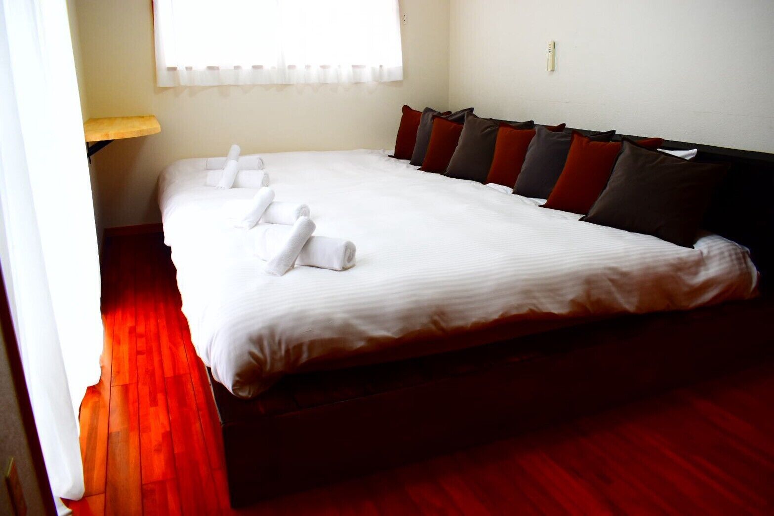 A neatly made bed with white linens and rolled towels, a wooden floor, and a window with white curtains.