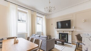 A living room with a fireplace, a television mounted above it, a dining table with chairs, and a sofa. - Elegant South Kensington Retreat (London)