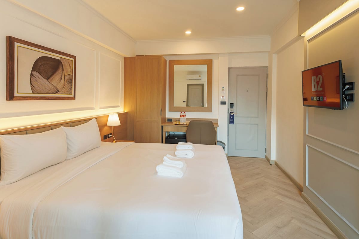A hotel room with a bed, bedside lamps, a desk with a chair, a mirror, a television, and a door.