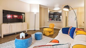 A living room with a TV displaying 'NETFLIX,' a sofa, ottomans, a floor lamp, and a kitchen in the background. - Fable 2Bd-2Ba MarinaDelRey Parking, Pool & Jacuzzi (Marina del Rey)
