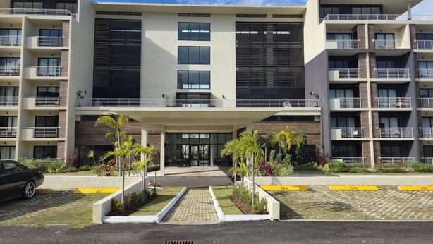 A modern multi-story building with balconies, a covered entrance, and a landscaped front yard.