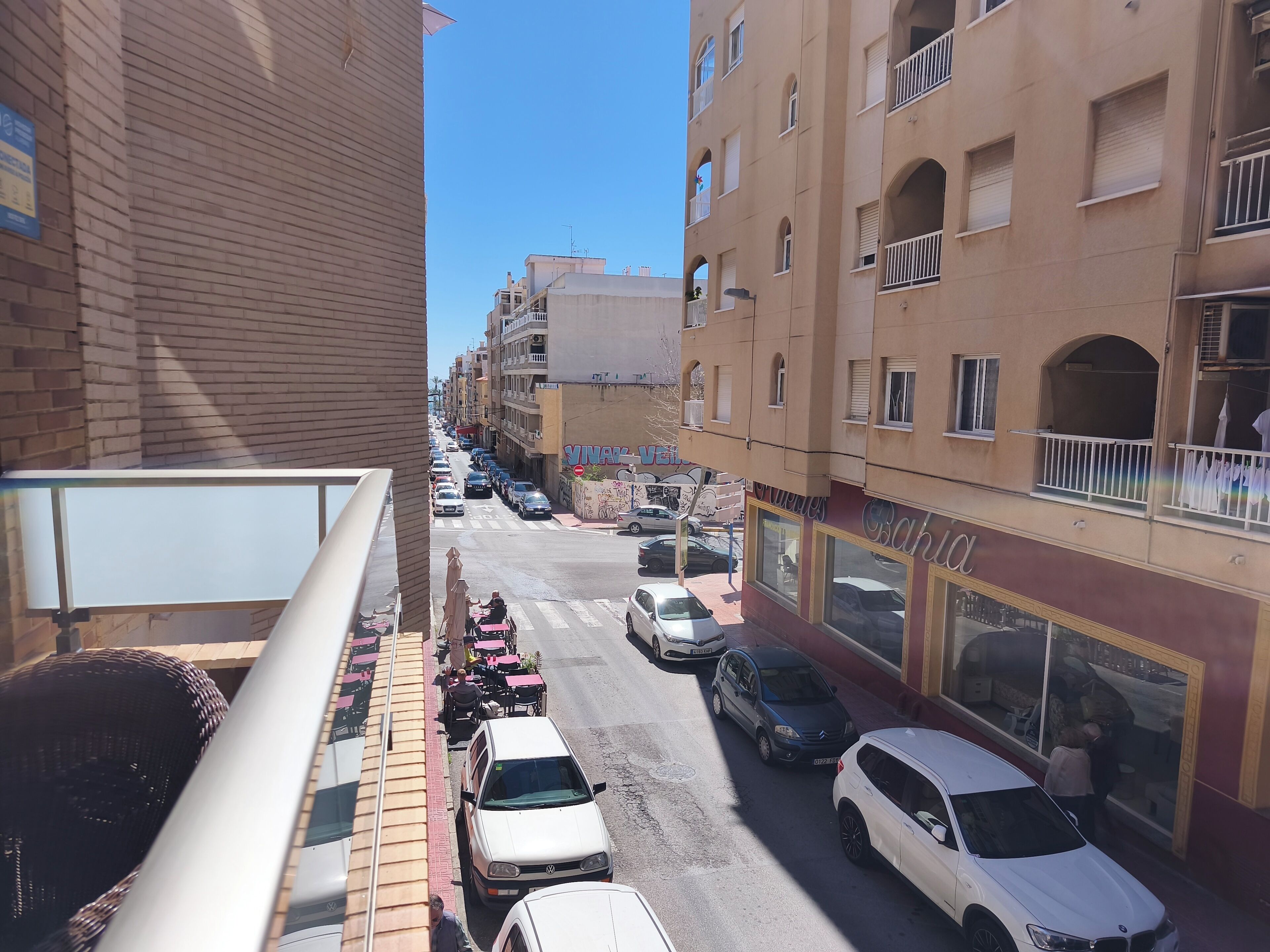 A street view with parked cars, a building with a storefront, and a balcony overlooking the street.