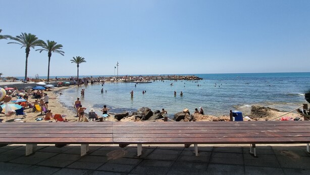 Ein Strand mit badenden und sonnenden Menschen, Palmen und einem Steg, der ins Wasser führt.