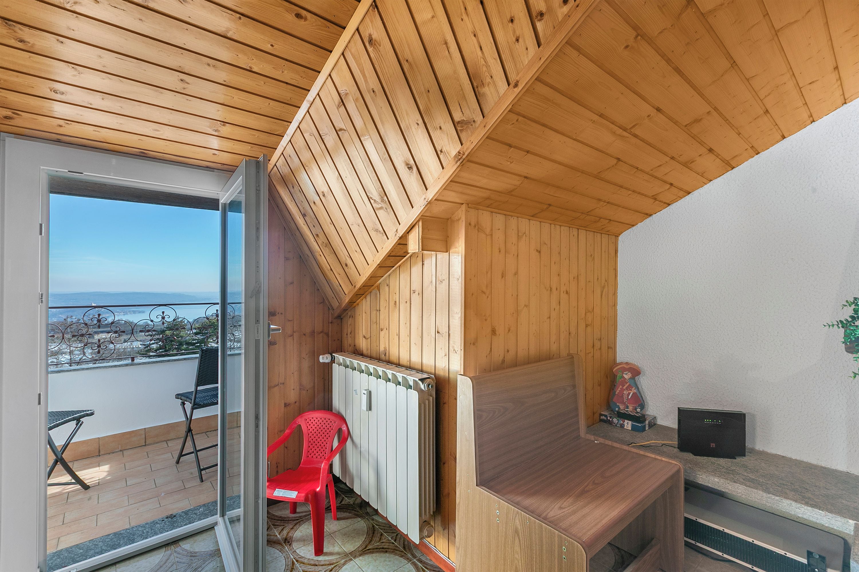 Un intérieur en bois avec une porte-fenêtre coulissante donnant sur un balcon, une chaise en plastique rouge et un radiateur.