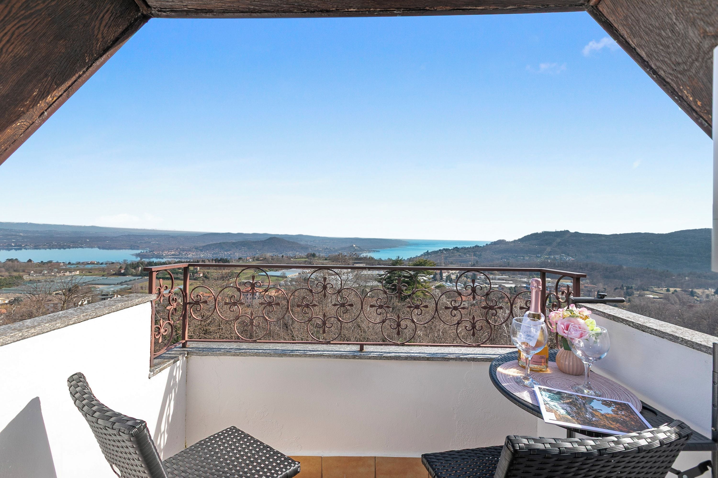 Un balcon avec vue sur un lac et des collines, une table avec une bouteille et des fleurs, et des meubles en osier.