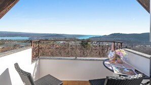 Un balcone con vista su un lago e delle colline, un tavolo con una bottiglia e fiori, e mobili in vimini.