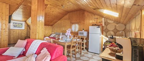 Interior acolhedor de uma cabana de troncos, com um sofá vermelho, mesa de jantar de madeira e cozinha equipada com micro-ondas e frigorífico.