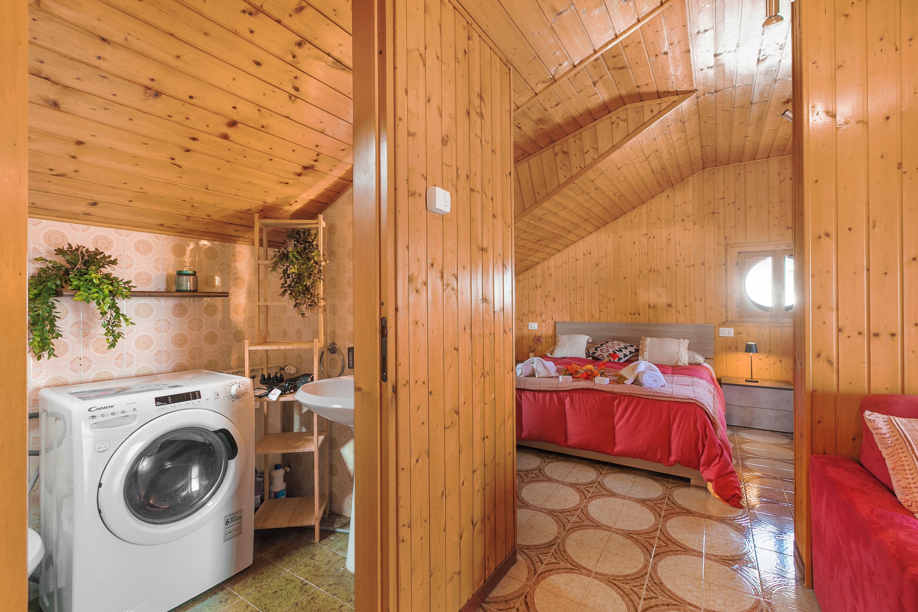 A wooden interior with a washing machine, a bed with red bedding, and a circular patterned floor.