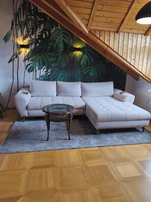 A living room with a sectional sofa, a round coffee table, a large wall mural, wooden flooring, and a sloped ceiling. - House 113 - Burgzauber Vacation Rental, Burghausen, 24-Hour Self-Check-In (Burghausen)