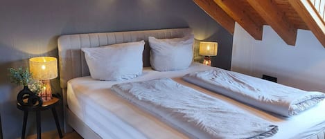 Ein Schlafzimmer mit schrägen Wänden, einem Bett mit weißer Bettwäsche, einem Nachttisch mit Lampe und einem kleinen Tisch mit Pflanze.
