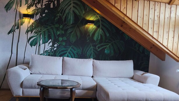 A room with a sectional sofa, a glass coffee table, a wooden herringbone floor, and a large wall mural of tropical foliage. - Vacation apartment Burgzauber (Burghausen)
