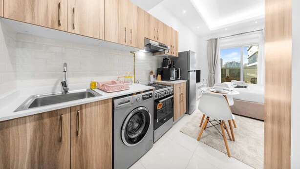 A modern kitchen with wooden cabinets, a stainless steel sink, a washing machine, and a dining area with a white table and chairs. - Free Housekeeping Visits During Your Stay - Stayshort - Cozy Studio in Sposrt City That Sleeps 3 (Dubai)