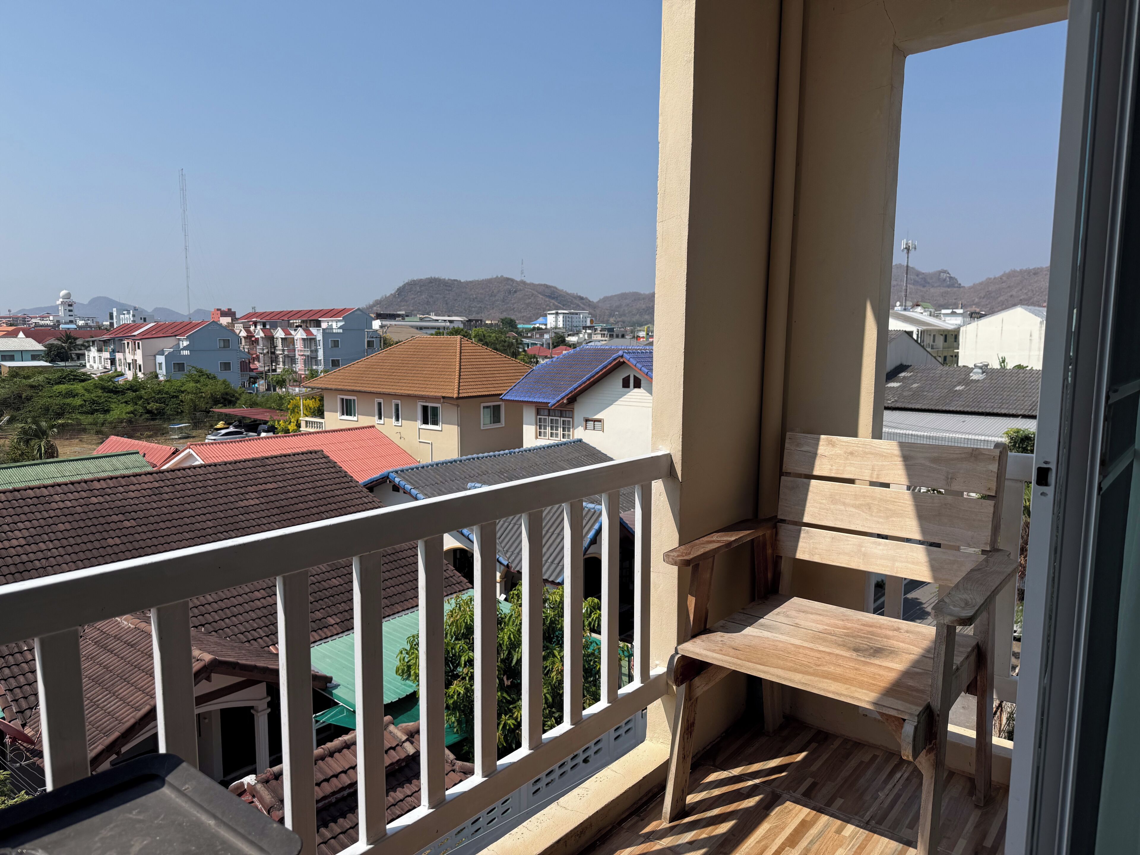 A balcony with a wooden bench, a view of a residential area with houses and a mountain in the background.