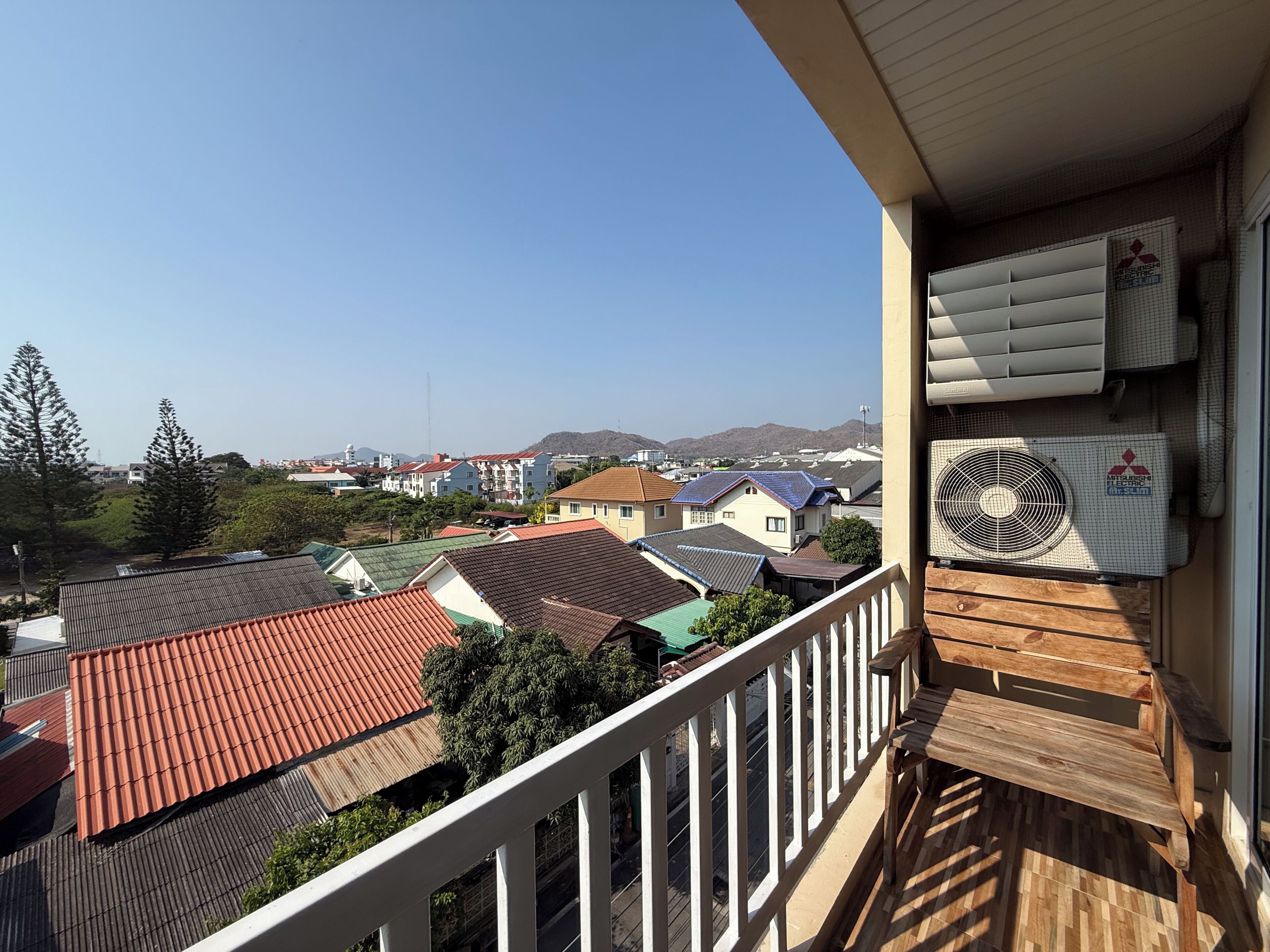 A balcony with a view of residential buildings, a Mitsubishi Electric air conditioning unit, and a wooden deck.