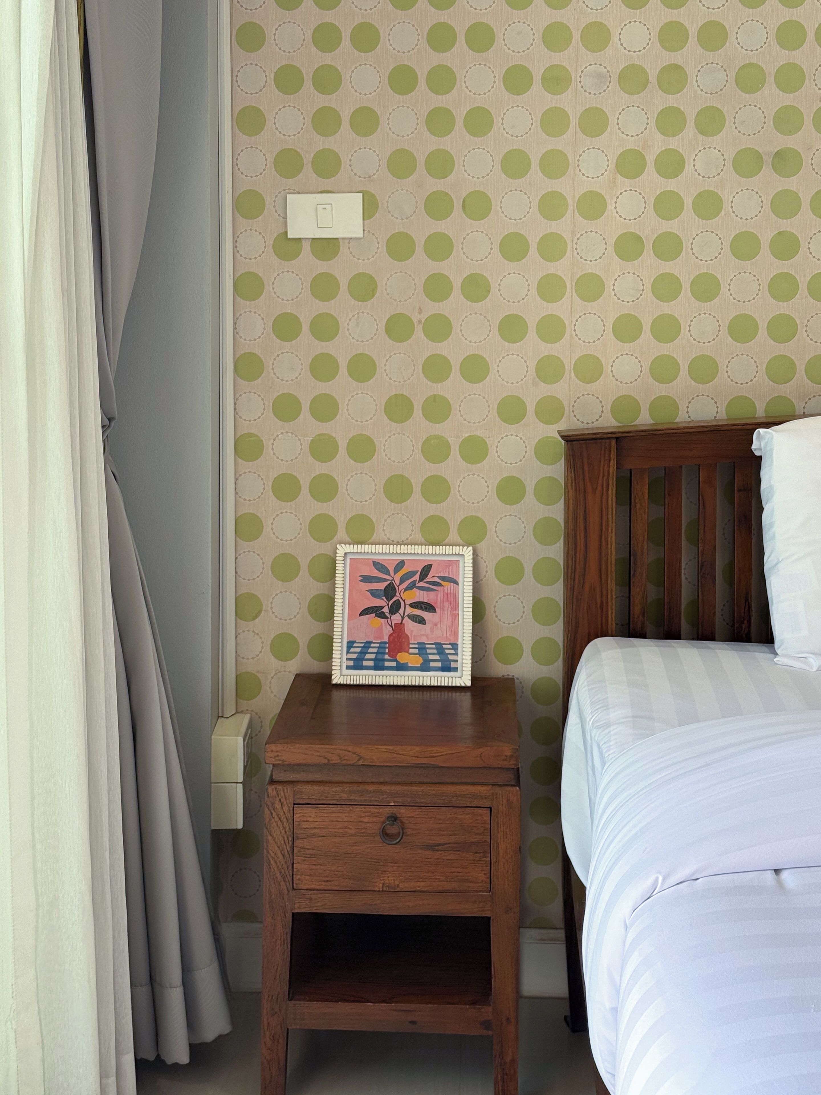 Un dormitorio con una mesita de noche de madera, una cama con ropa de cama blanca y un papel tapiz verde con lunares.