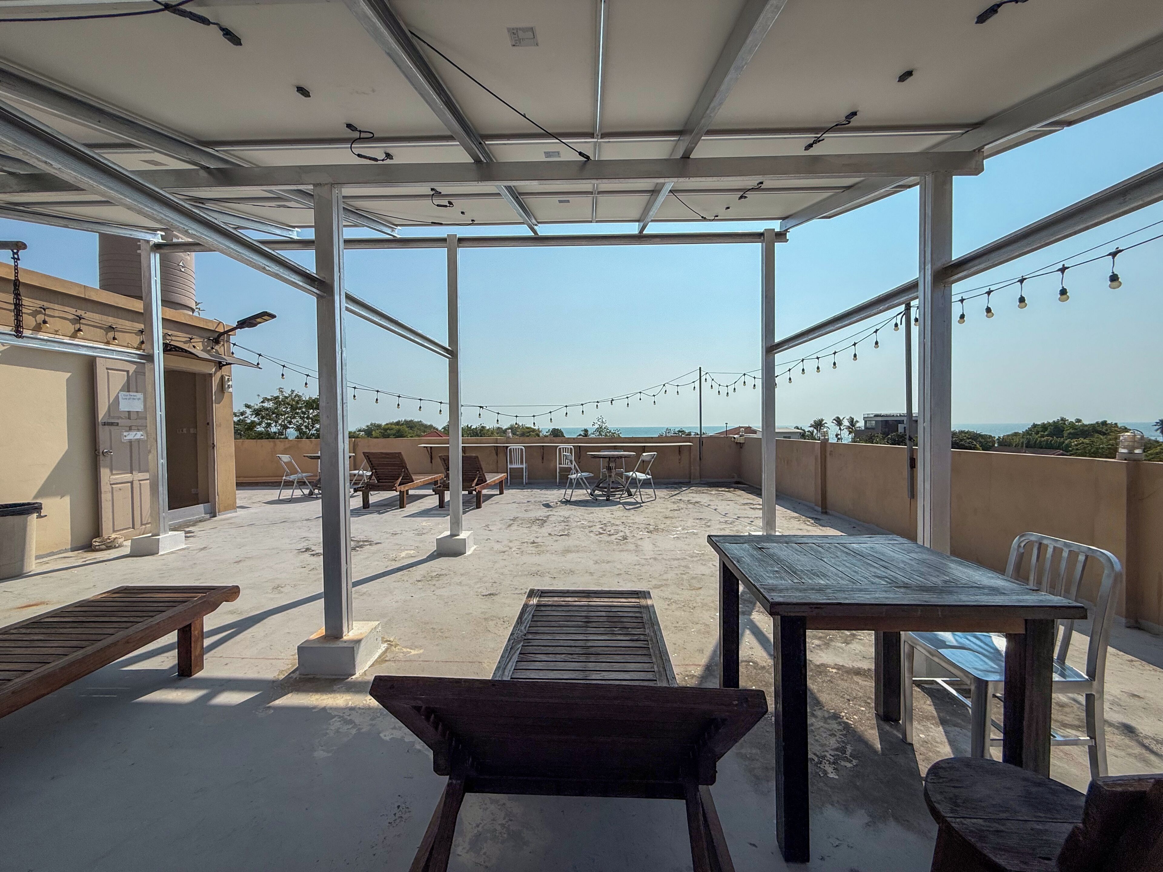 A rooftop terrace with a view of the sea, featuring wooden furniture and string lights.