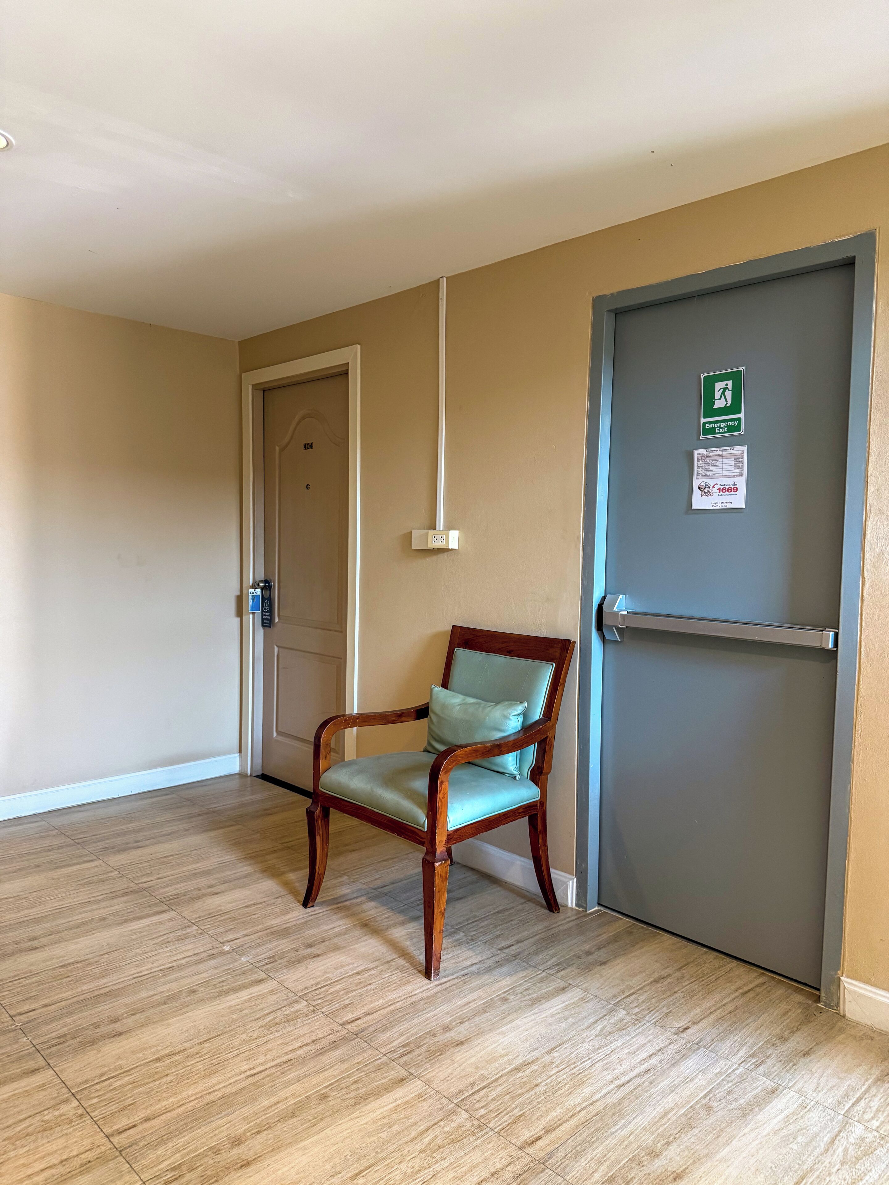 Una stanza con una sedia di legno, una porta con il segnale di uscita di emergenza e una presa elettrica fissata al muro.