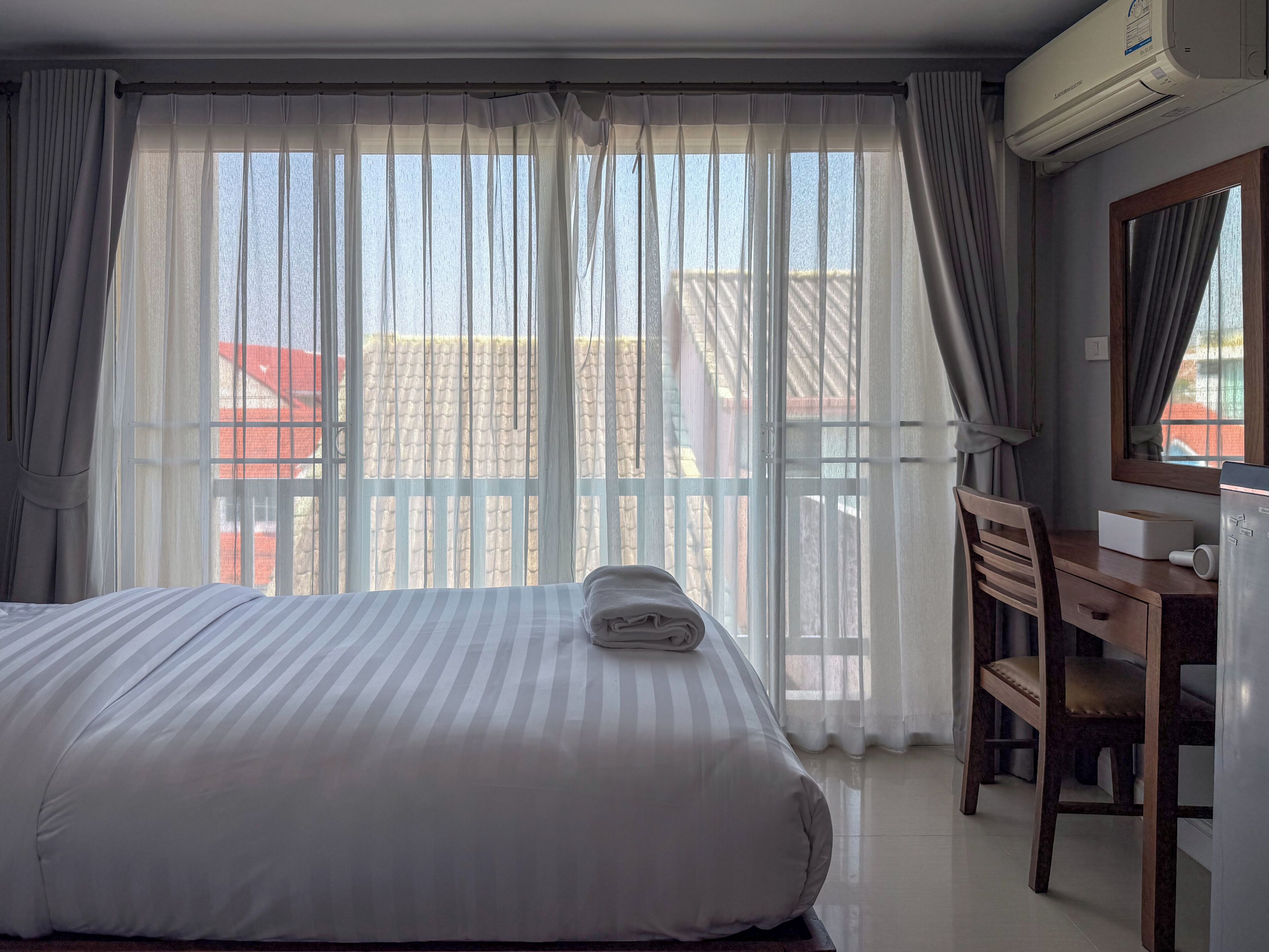 Habitación de hotel con cama, escritorio, aire acondicionado y vistas a edificios a través de cortinas traslúcidas.
