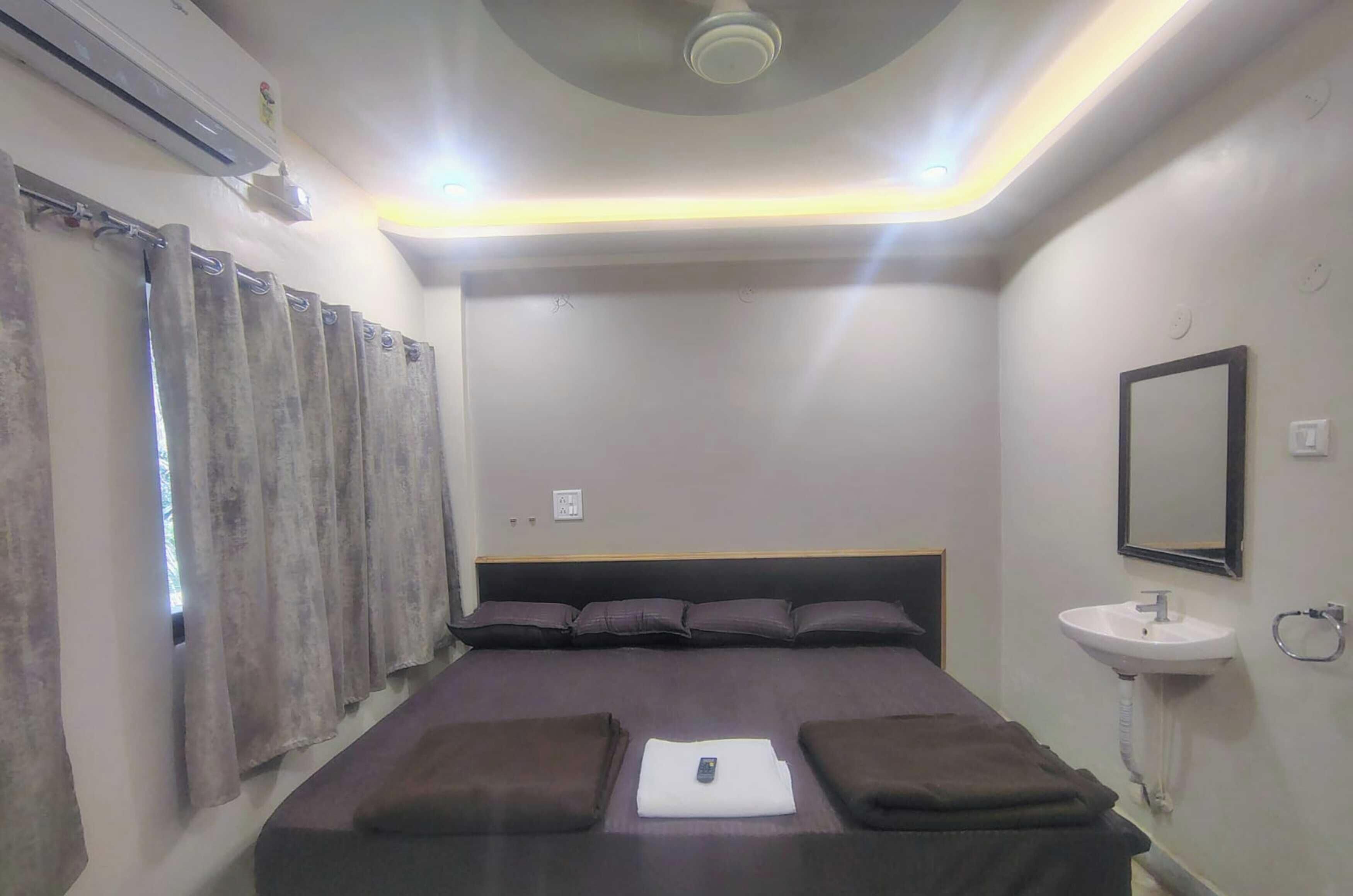 A hotel room with a bed, curtains, a ceiling fan, a bedside table, and a wall-mounted sink.