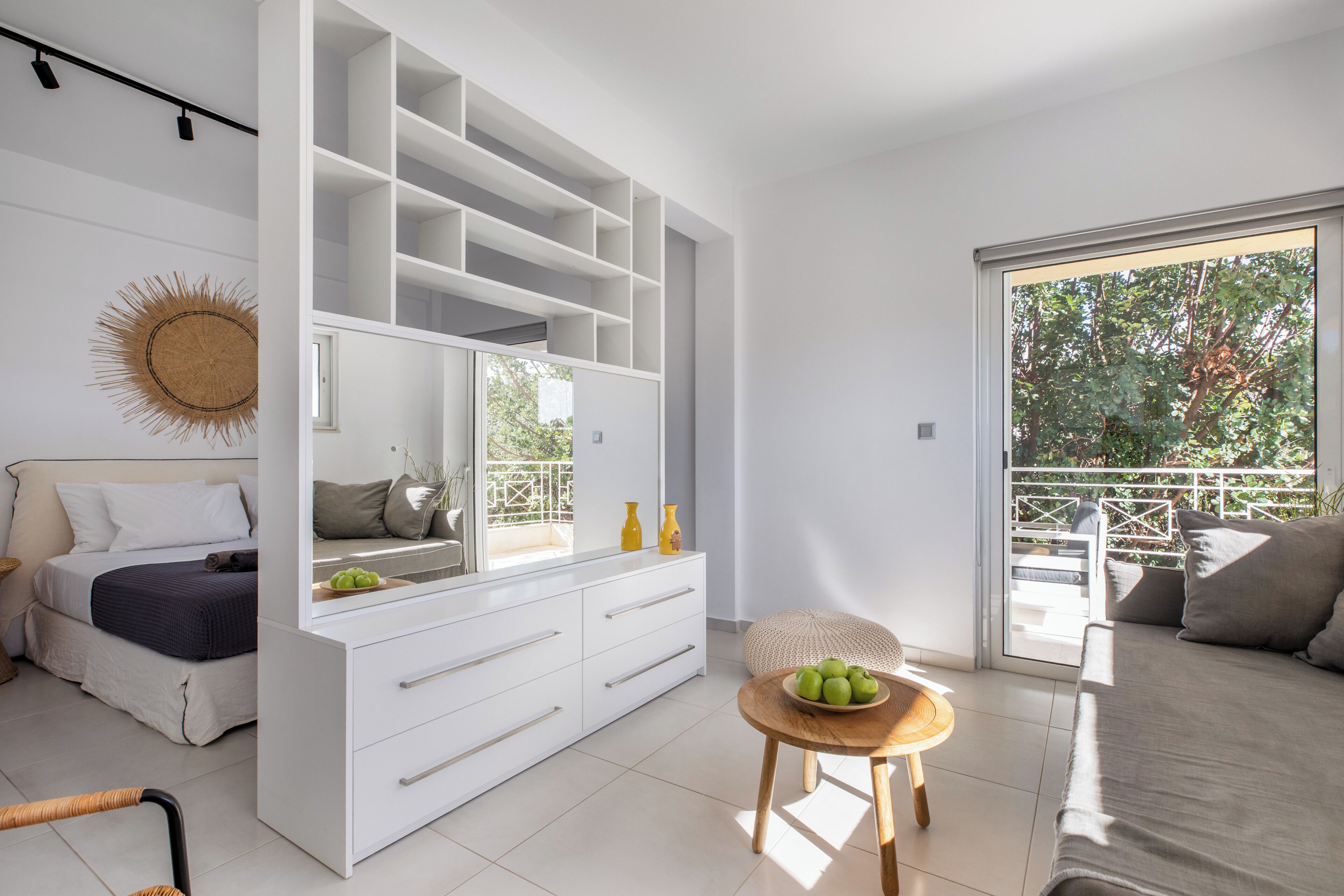A modern bedroom with a large bed, a built-in wardrobe, a small round table with a bowl of fruit, and a balcony with a view of trees.