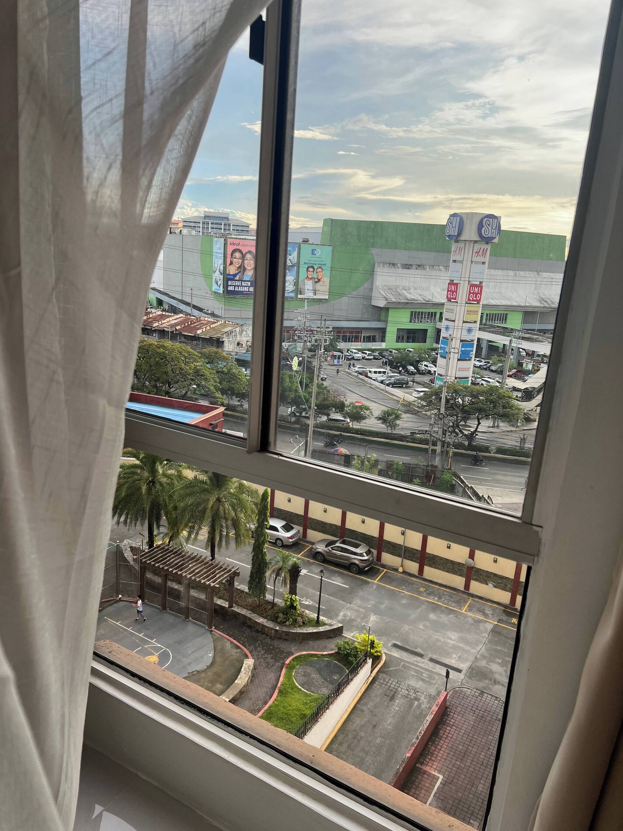A view from a window with a curtain, overlooking a parking lot, a green building with multiple signs, and a street with vehicles.
