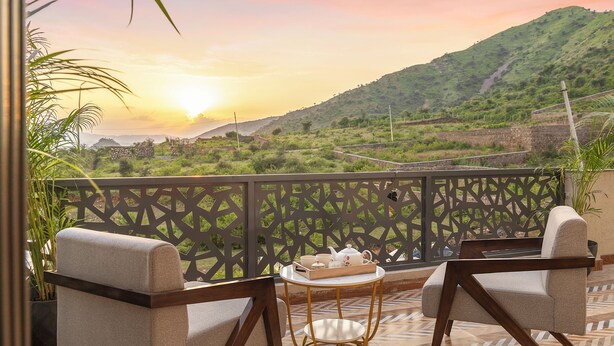 A balcony with a view of a hilly landscape, a table with a tray of items, and two chairs. - ELIVAAS Shreya Blossom 4 BHK With Pool (Udaipur)