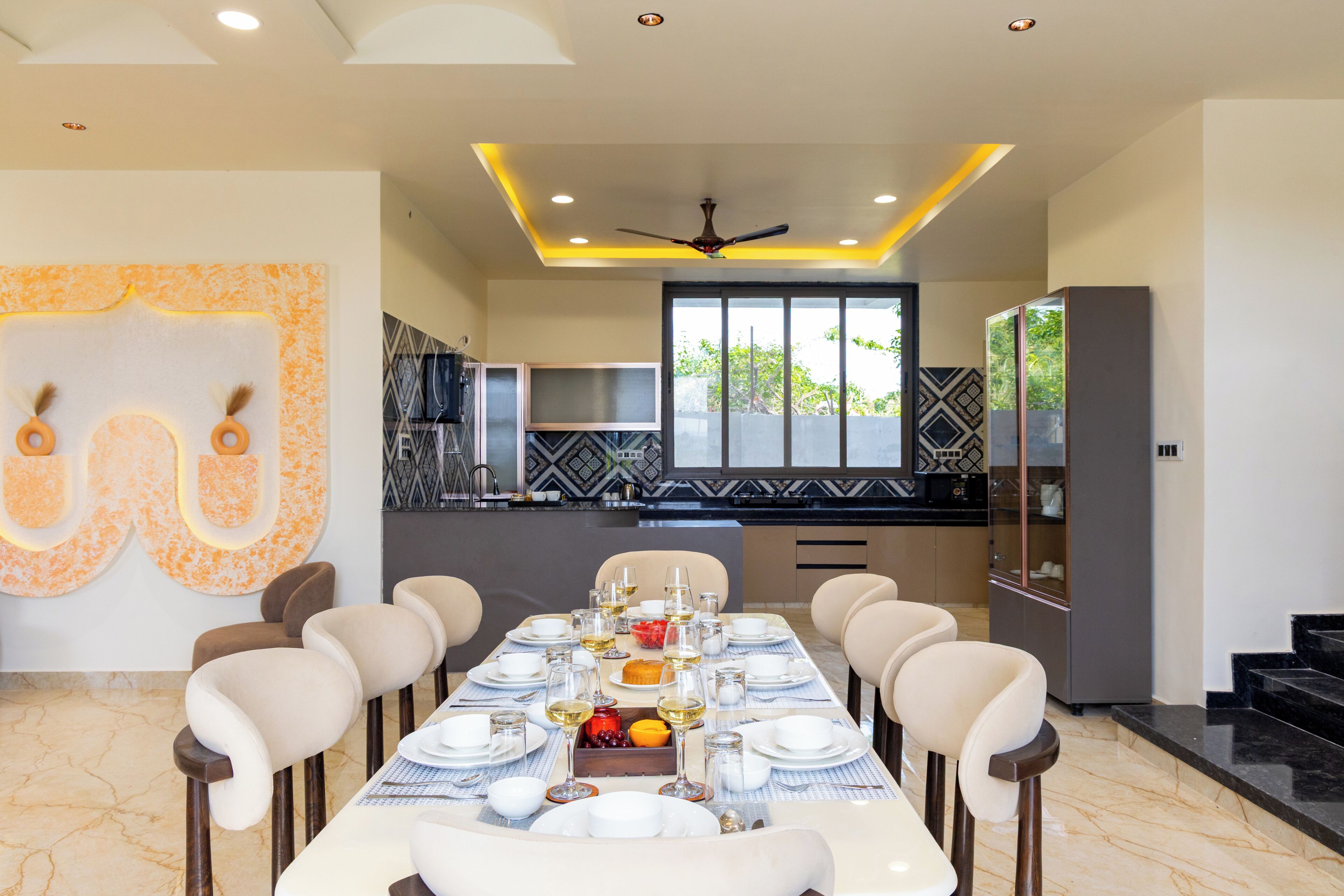 A dining area with a long table set for six, a ceiling fan, and a kitchen in the background.