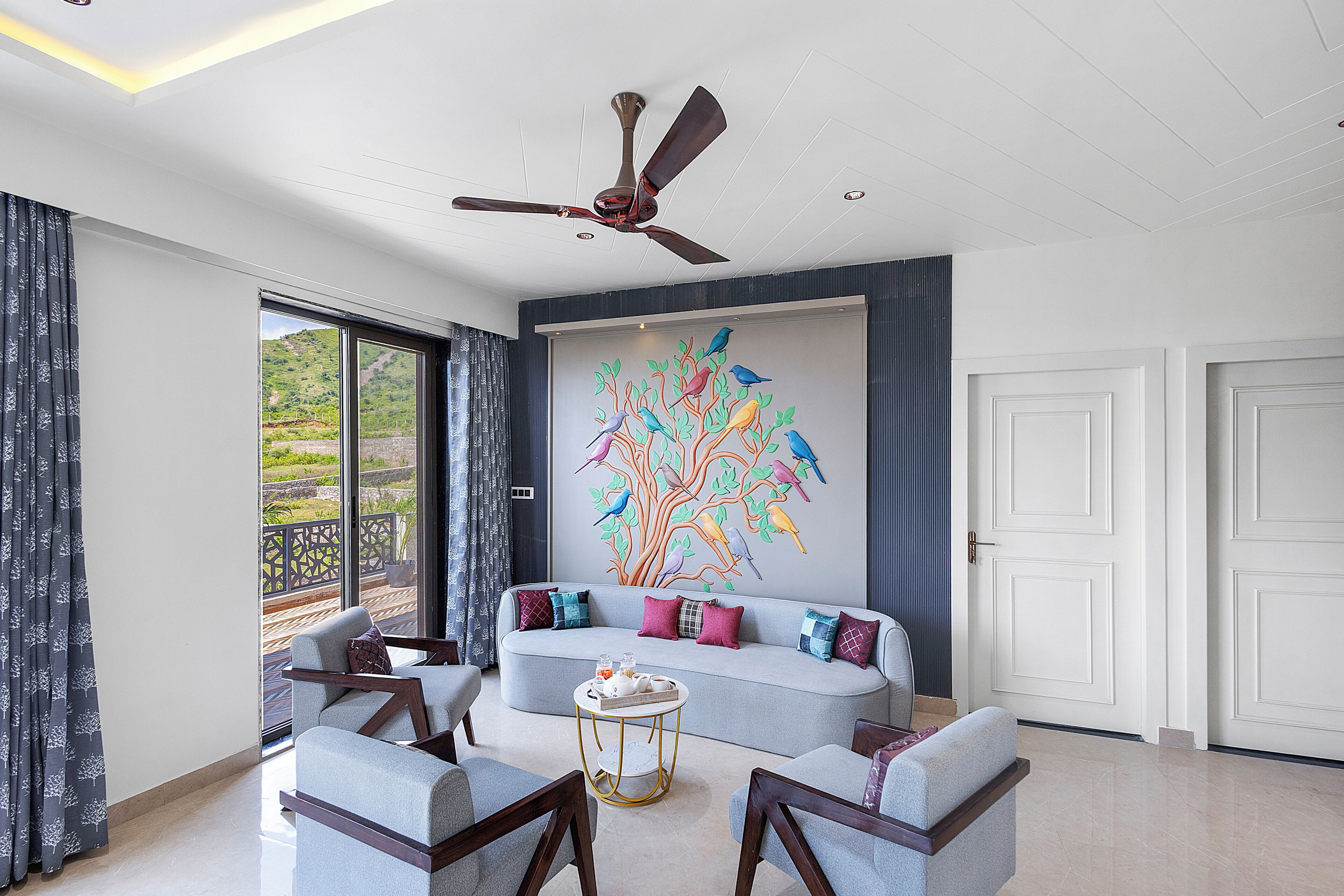 A modern living room with a sofa, chairs, a ceiling fan, and a large window with a view.