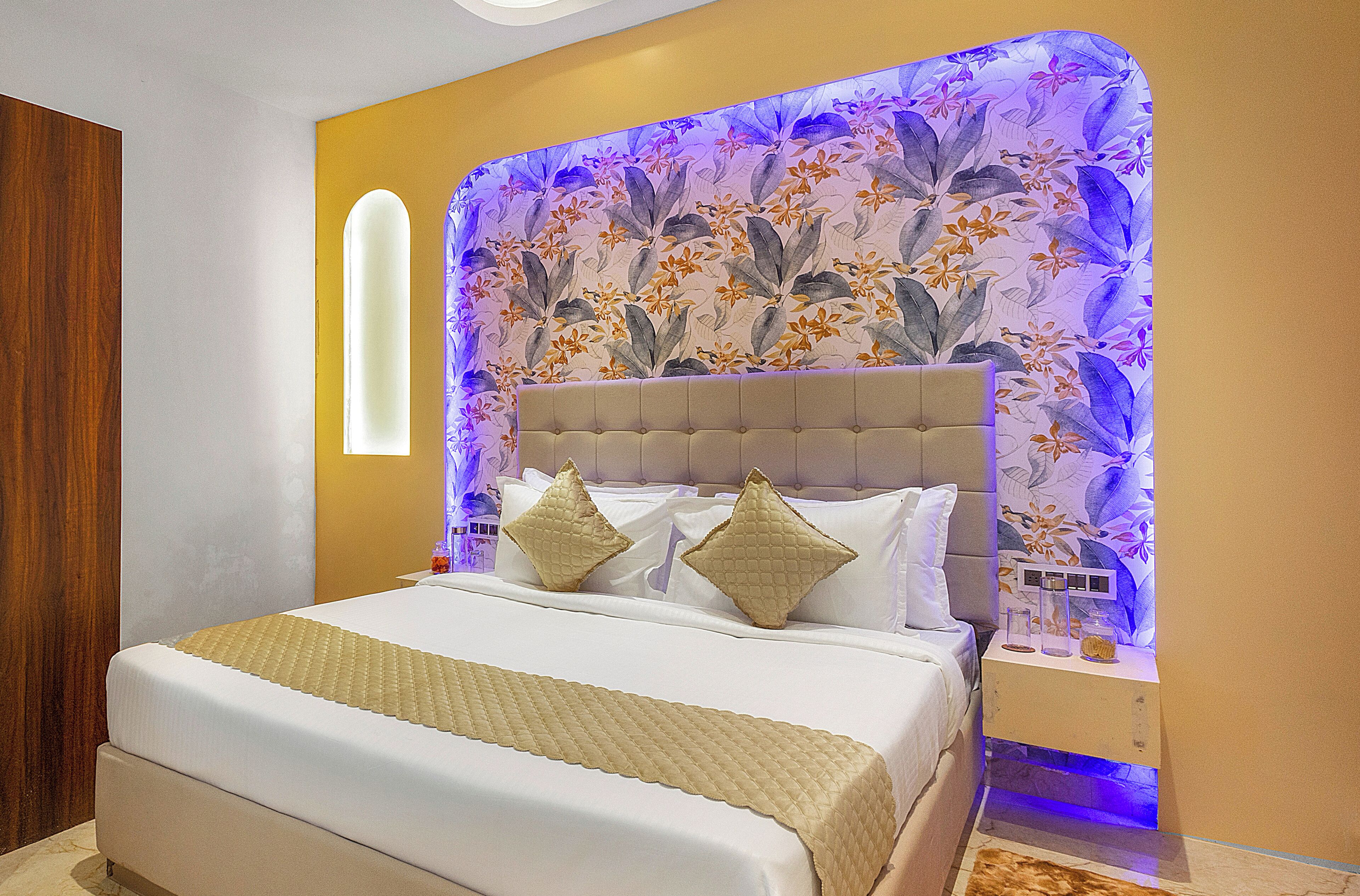 A bedroom with a bed, a headboard with floral wallpaper, and a bedside table with a lamp.