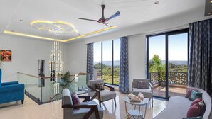 A modern living room with a ceiling fan, large windows, and a balcony with a view. - ELIVAAS Shreya Blossom 4 BHK With Pool (Udaipur)