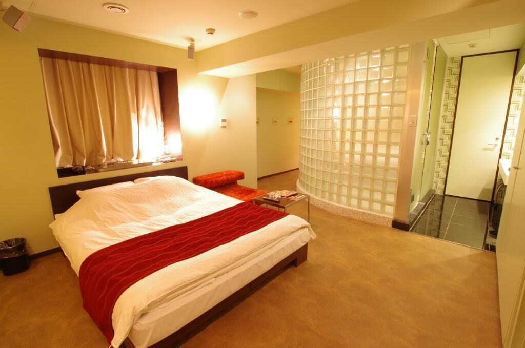 A hotel room with a bed, a small table, a window with curtains, a shower area, and a bathroom with a glass door.